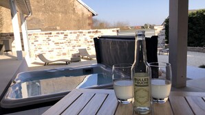 Una mesa de madera con una botella de BULLES DE RUCHES y dos vasos de líquido blanco, presumiblemente una bebida, en un entorno exterior con piscina y sillones de relax.