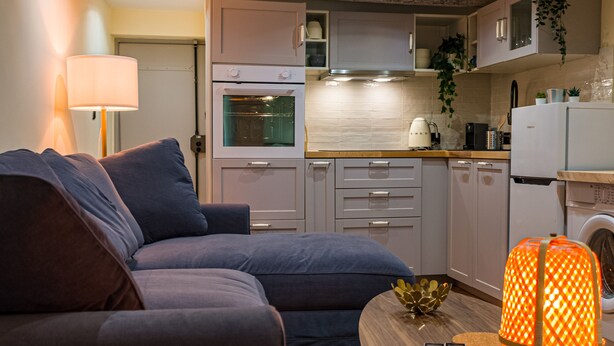 A compact living space with a sofa, a wooden coffee table, a lamp, and a kitchen area including a stove, oven, and refrigerator. - Cosy new apartment (Nice)