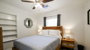 Ein Schlafzimmer mit einem hölzernen Bett, weißen Wänden, einem Deckenventilator, einem Fenster mit Vorhängen, einem Nachttisch mit Lampe und einem Spiegel an der Wand.