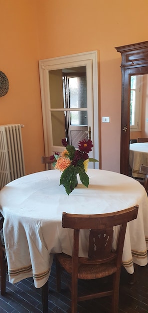 Un coin repas avec une table ronde recouverte d’une nappe blanche, un vase de fleurs et une chaise en bois.