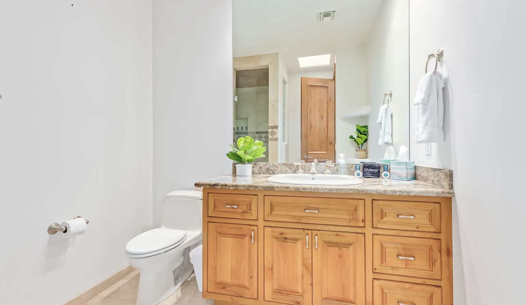 Une salle de bain avec des toilettes blanches, des meubles en bois, un comptoir en granit, un lavabo, un miroir et une plante en pot.