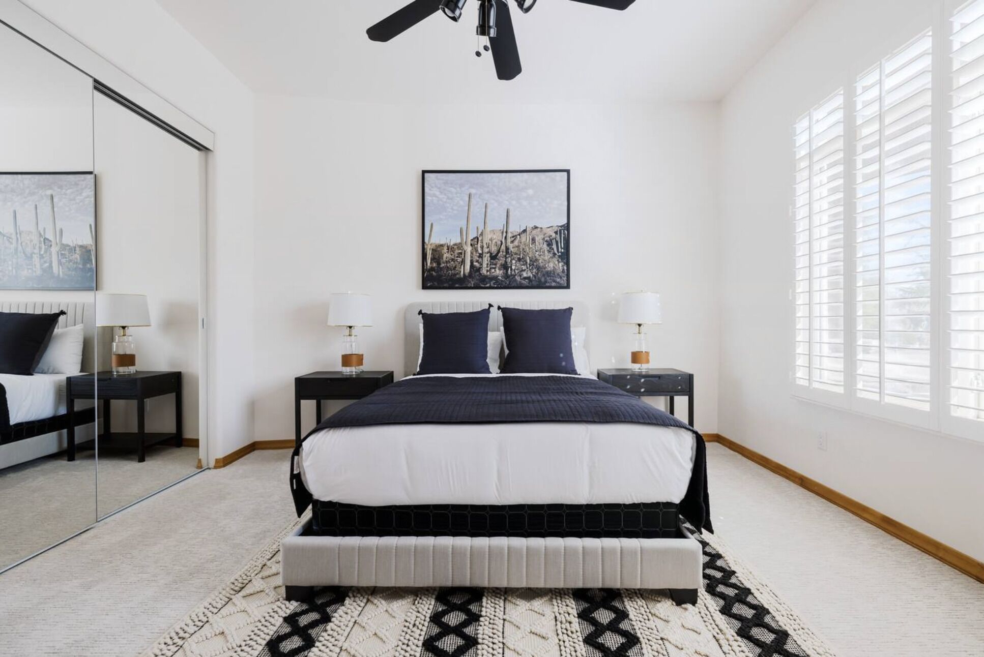A bedroom with a bed, two bedside tables, a ceiling fan, a large window with blinds, and a wall-mounted picture.