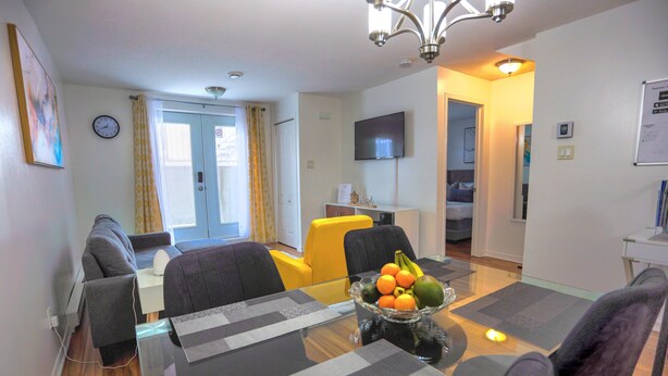 Un comedor con una mesa de vidrio, individuales grises y negros, y un frutero. Hay dos sofás grises y una silla amarilla. Un televisor de pantalla plana está montado en la pared, y una puerta conduce a otra habitación.