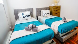 Habitación de hotel con dos camas, cada una con cabecero gris y colcha verde azulada. Hay dos almohadas con texto, un aparador de madera y un pequeño florero encima.