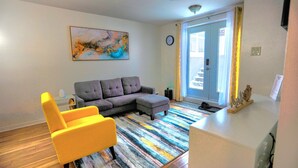 Una sala de estar con sofá, un sillón amarillo, un tapete colorido a rayas, un reloj y una puerta con cortinas.