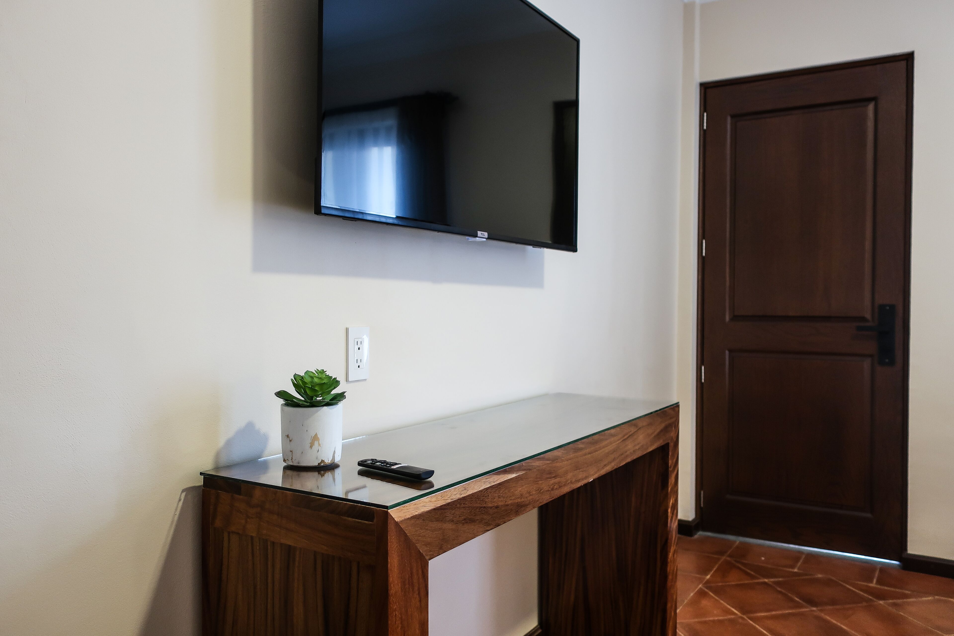 Una habitación moderna con una televisión de pantalla plana montada en la pared, un escritorio de madera con una planta en maceta y una puerta de madera cerrada.