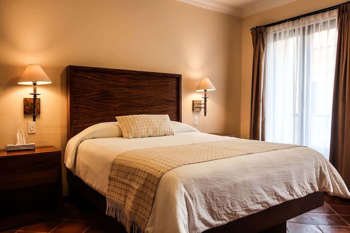 A neatly made bed with a wooden headboard, two bedside lamps, and a window with curtains.