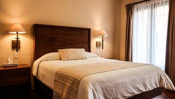 Una cama bien tendida con cabecera de madera, dos lámparas de noche y una ventana con cortinas.