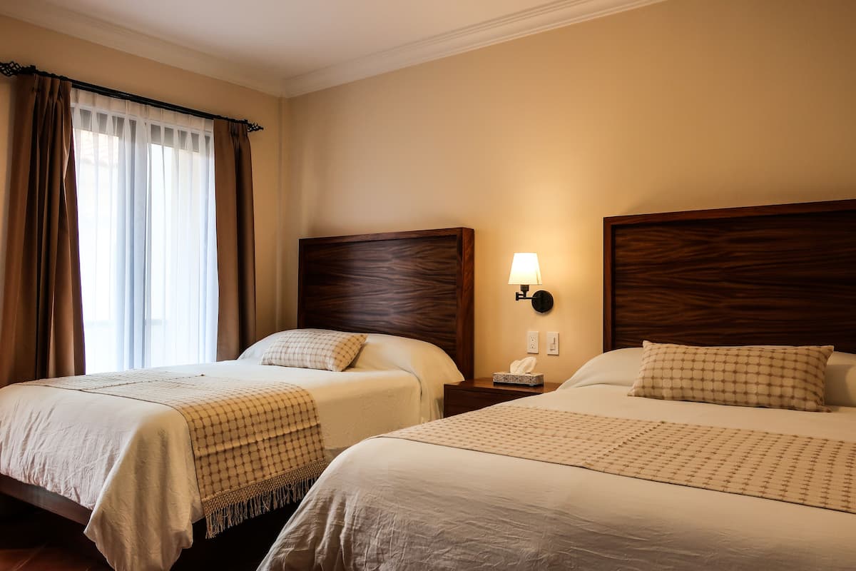 A hotel room with two beds, each with a wooden headboard, a bedside table with a lamp, and a window with curtains.