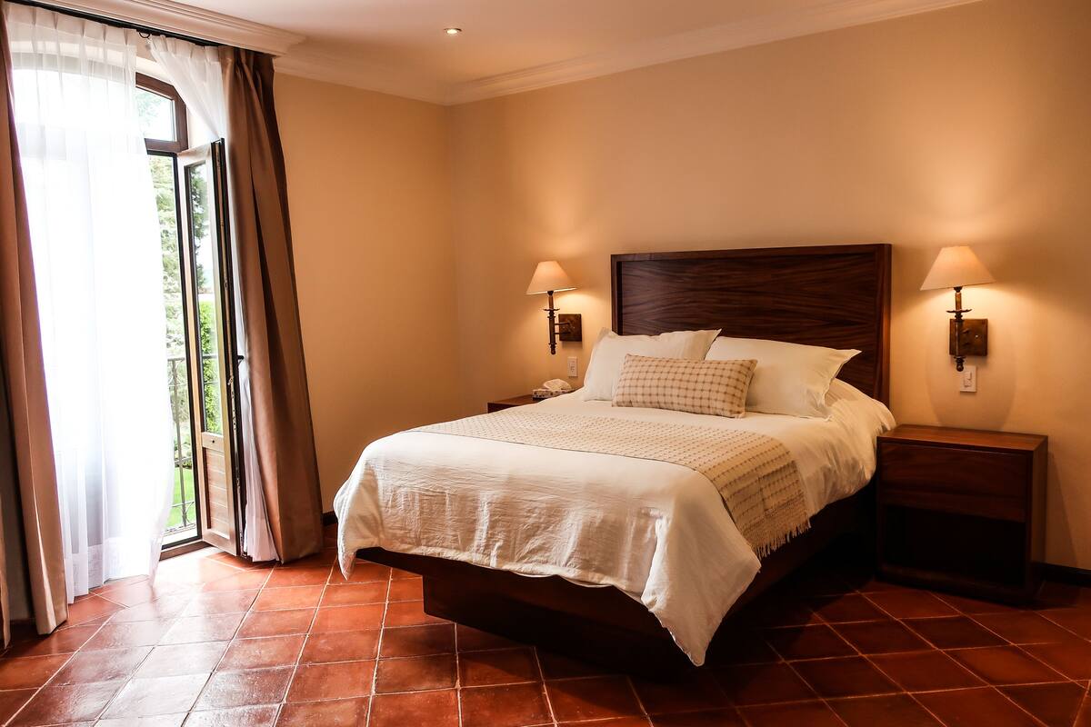 A neatly made bed with a wooden headboard, two bedside lamps, and a view of a garden through a glass door.
