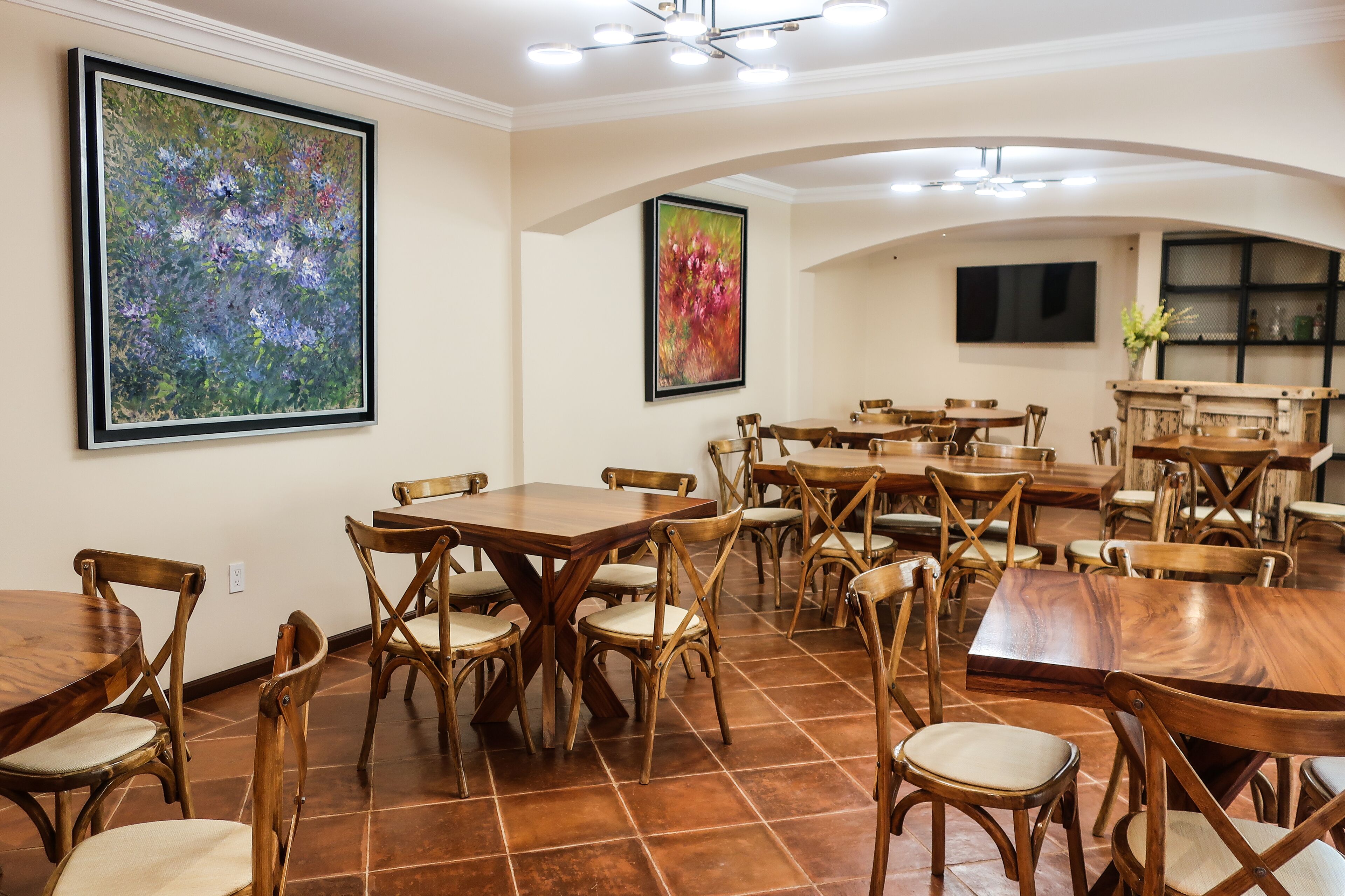 A dining area with multiple wooden tables and chairs, a large floral painting on the wall, and a television mounted on the wall.