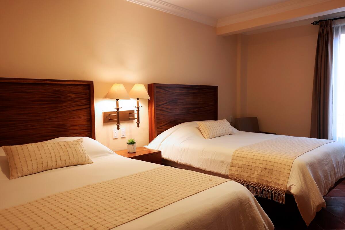 A hotel room with two beds, a nightstand with a lamp, and a window with curtains.