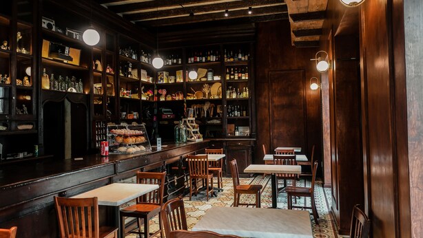 Un bar con estantes de madera llenos de botellas y diversos artículos. Hay varias mesas y sillas dispuestas para sentarse.