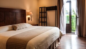 Un dormitorio con una cama grande, cabecera de madera, mesita de noche con lámpara, ventana con cortinas y vistas al exterior con vegetación.