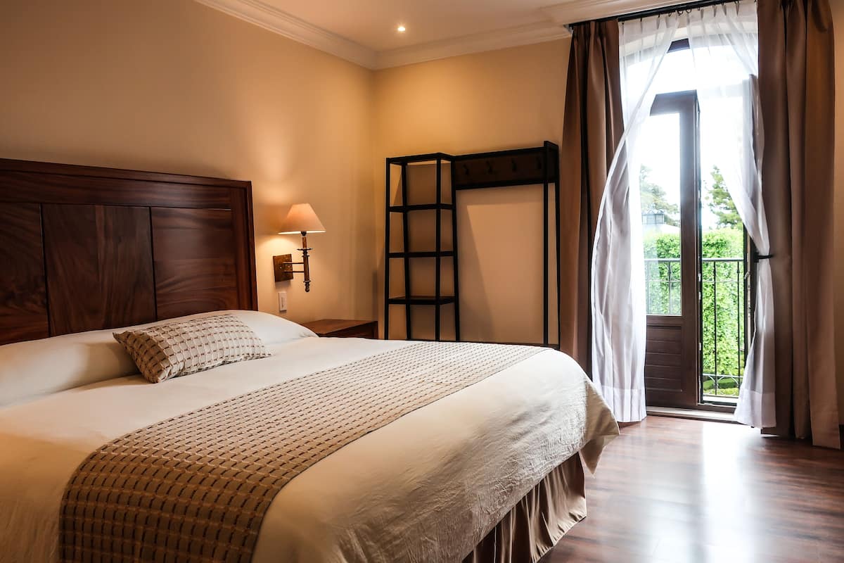 A bedroom with a large bed, a wooden headboard, a nightstand with a lamp, a window with curtains, and a view of greenery outside.