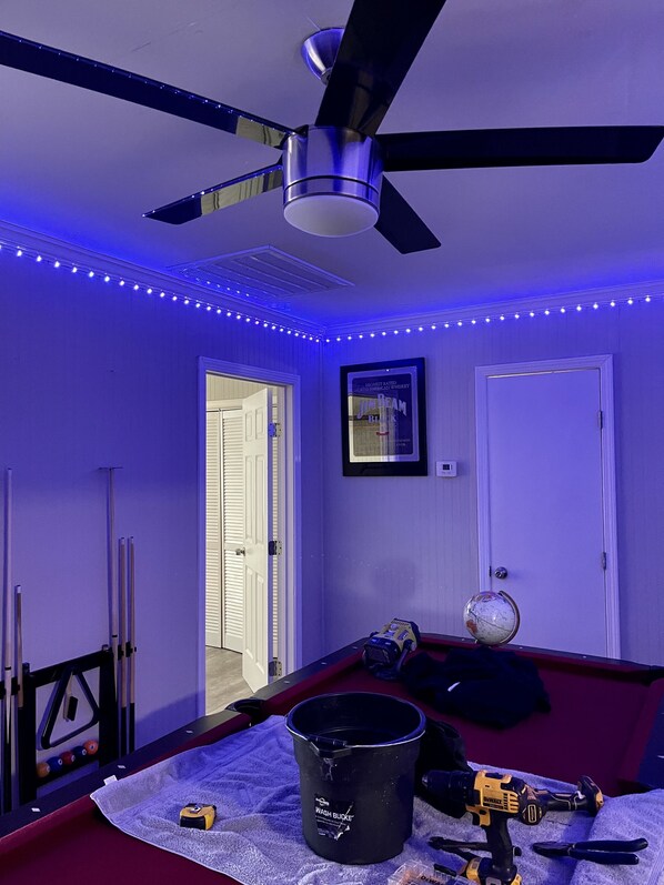 A room with a ceiling fan, LED strip lights, a framed picture, a globe, a bucket, and a power tool on a red couch.