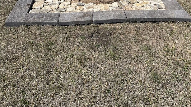 Un hoyo para fuego de piedra en un campo cubierto de hierba, con una caravana y una casa azul al fondo.