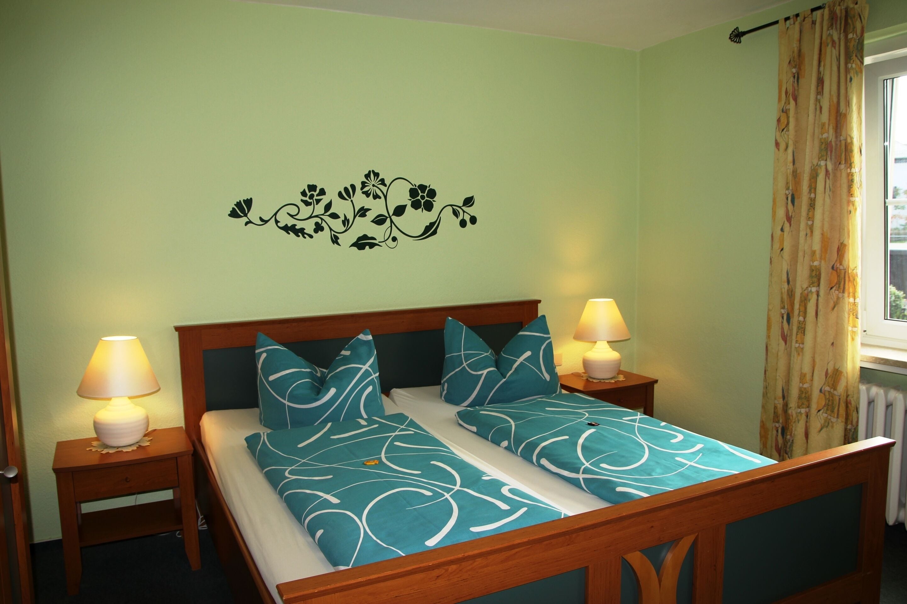 Una cama bien tendida con una colcha estampada en tonos aguamarina y blanco, flanqueada por dos lámparas de noche sobre mesitas de madera.