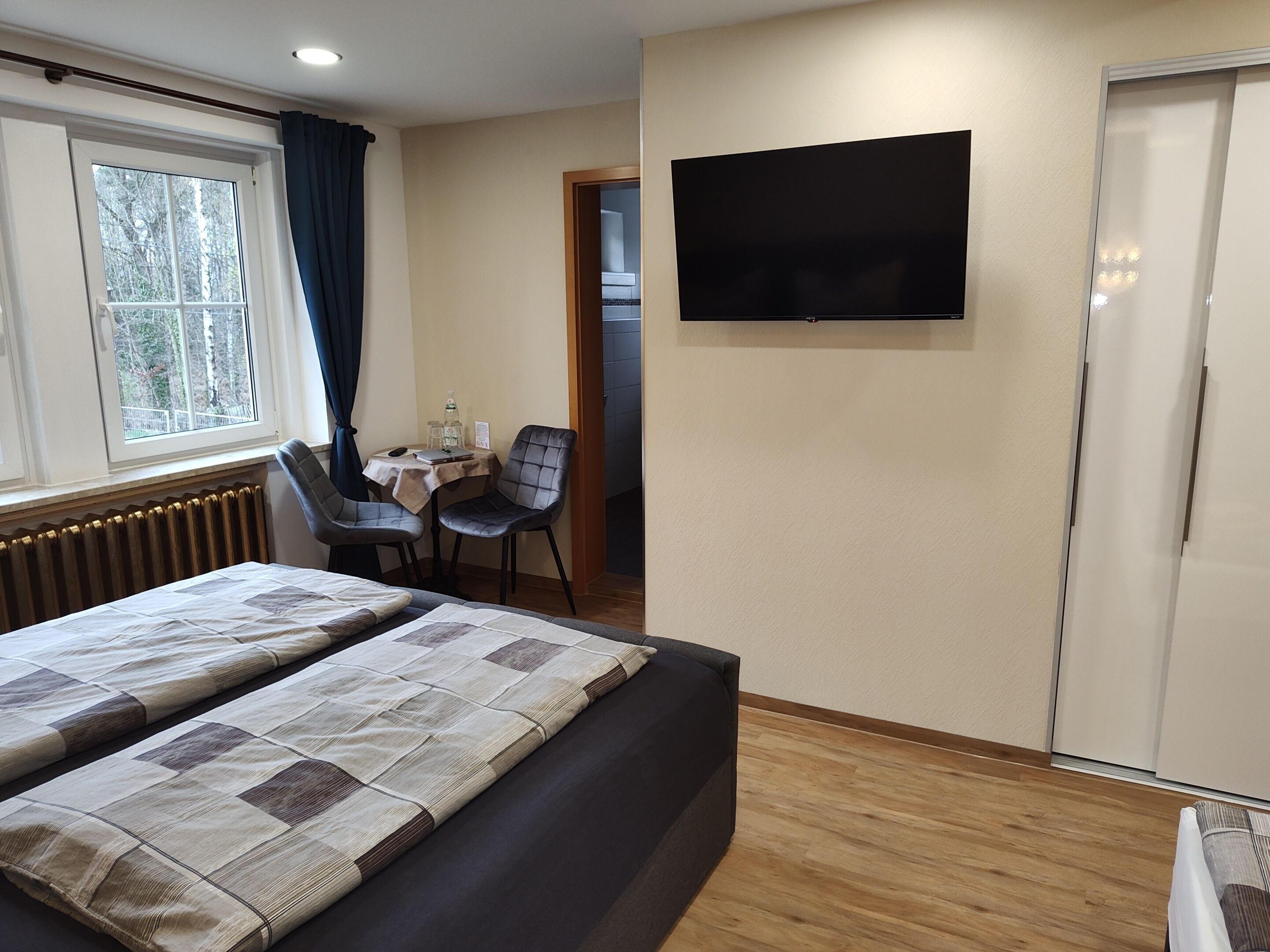 A hotel room with a bed, a desk with two chairs, a television mounted on the wall, and a window with curtains.