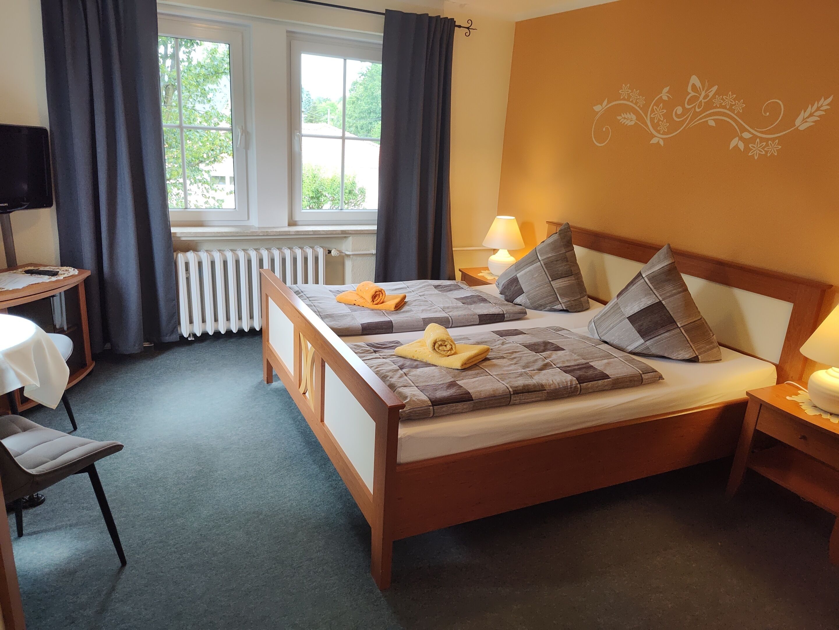 A hotel room with two beds, a table, a chair, a lamp, and a view of greenery through the window.