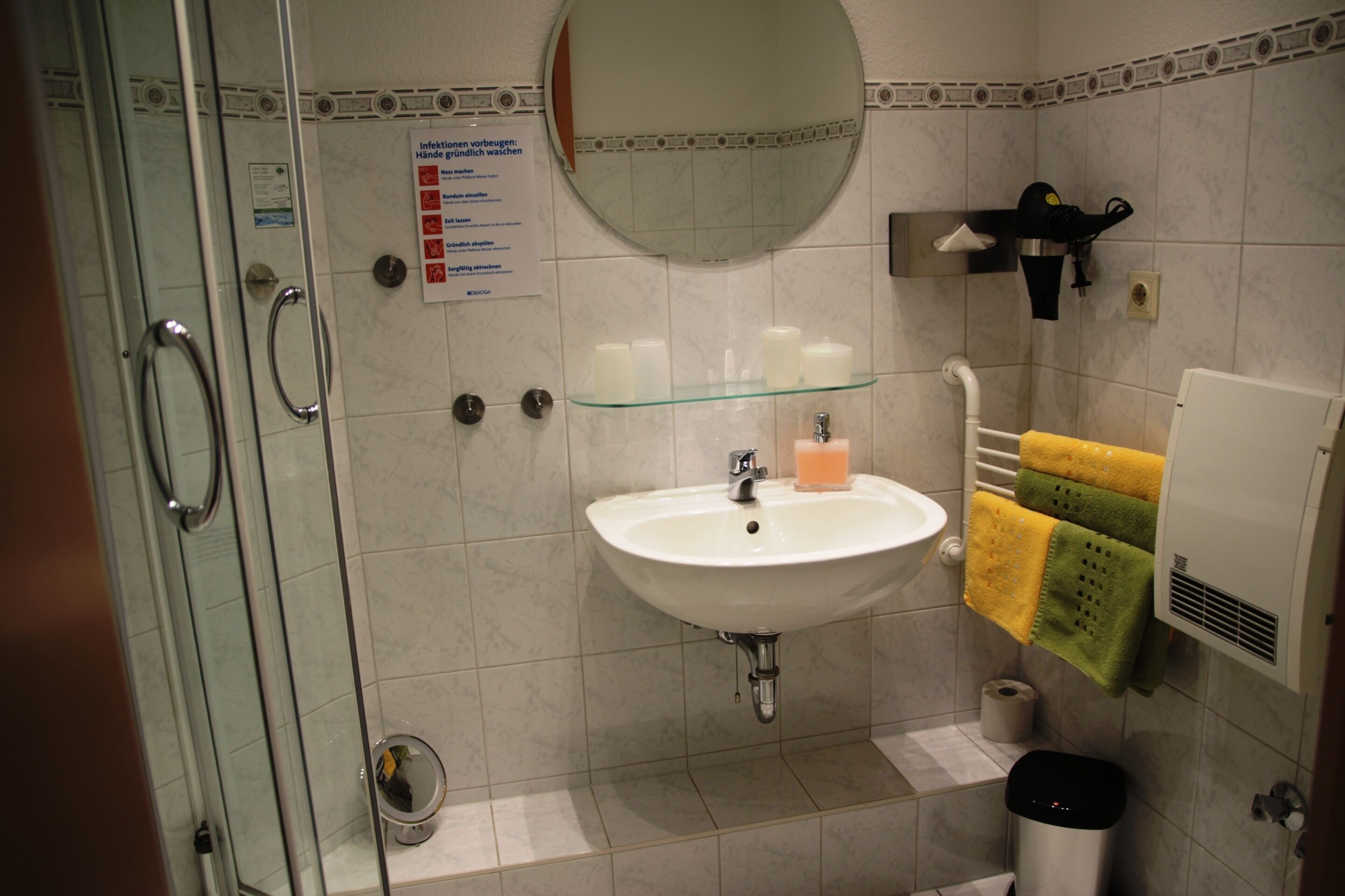A bathroom with a glass shower enclosure, a sink with a soap dispenser, and a towel rack with yellow and green towels.