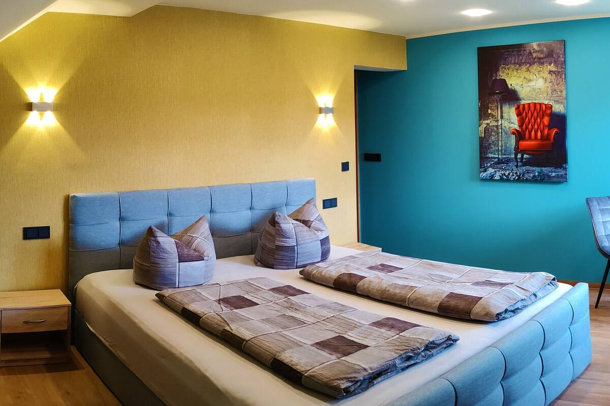 A bedroom with a large bed, two wall-mounted lights, a painting, and a chair.