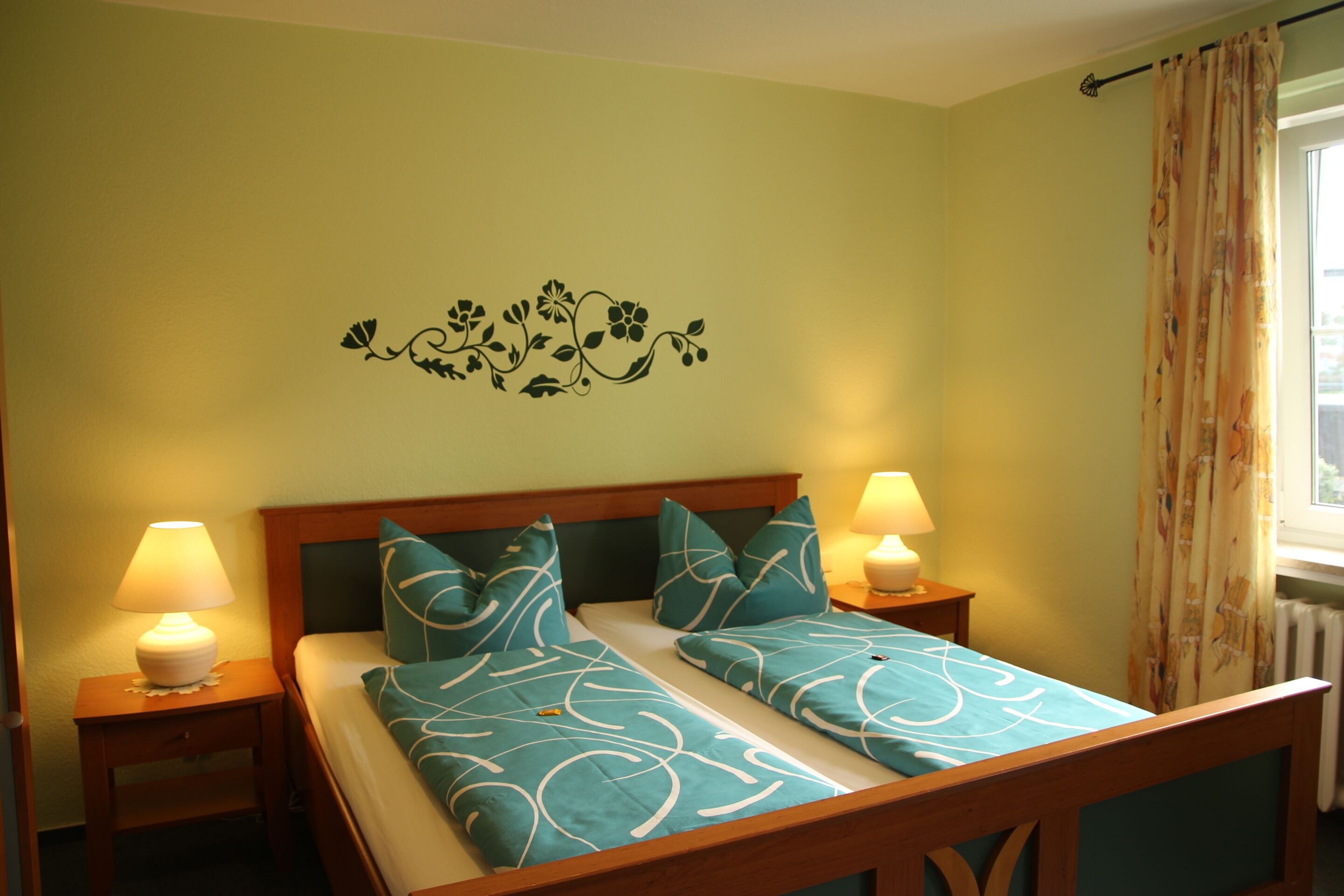 Een tweepersoonsbed met blauwgroene beddengoed en passende kussens, twee bedlampjes, een bloemmotief op de muur en een raam met gordijn.