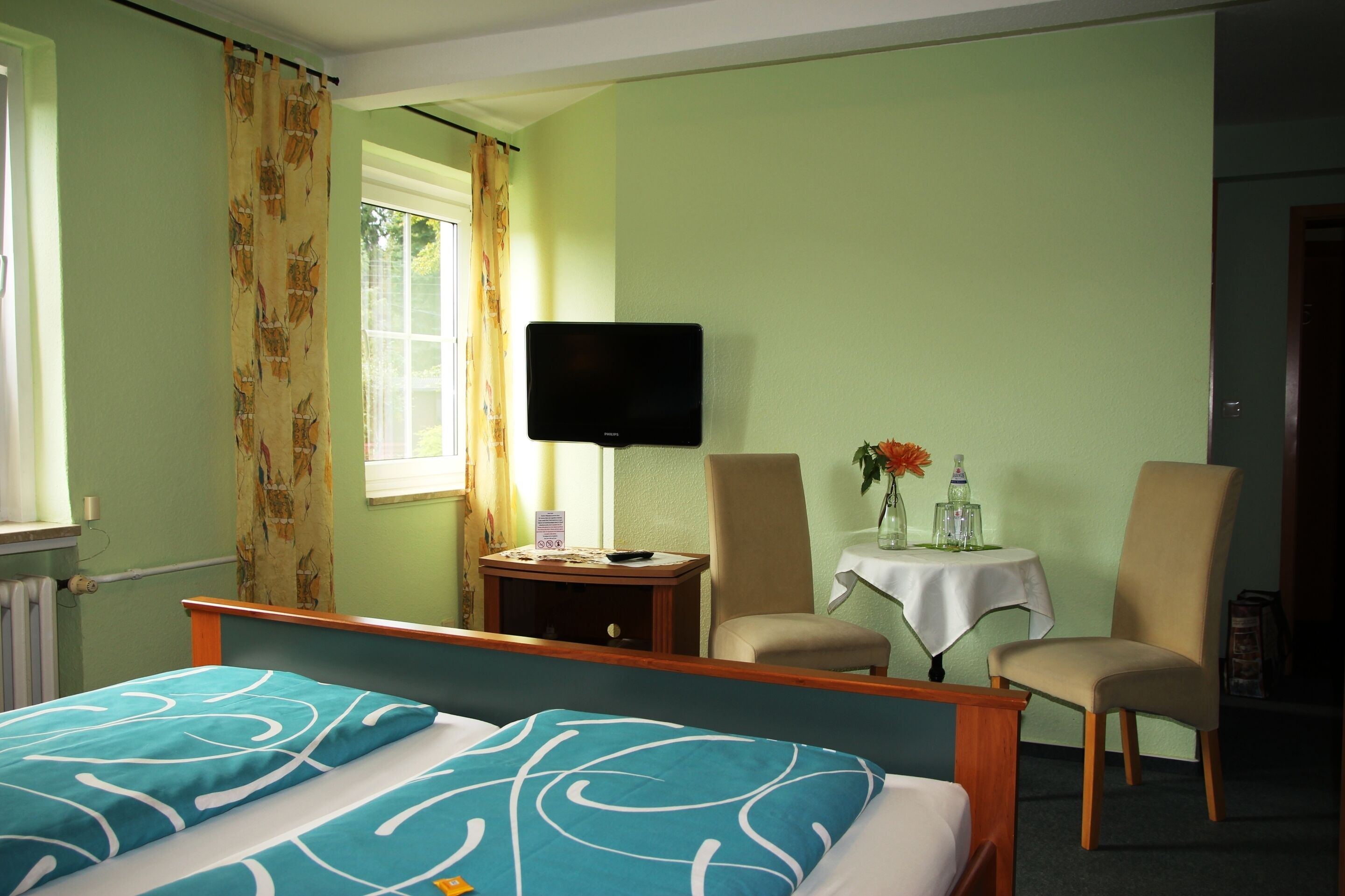 A hotel room with a bed, a television, two chairs, a small table with a vase, and a window with curtains.