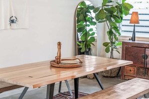 A wooden table with a woven tray and a candle holder, a bench, and a mirror with a plant in the background.