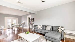 A spacious living room with a grey sectional sofa, a white coffee table, and a dining area with chairs and a table. - 2 BR • Oak Lawn House by Uptown • WFH Setups (Dallas)