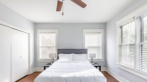 A bedroom with a bed, two bedside tables, a ceiling fan, and large windows with blinds. - 2 BR • Oak Lawn House by Uptown • WFH Setups (Dallas)