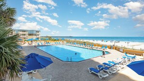A pool area with lounge chairs and umbrellas, a clear view of the beach, and a building in the background.