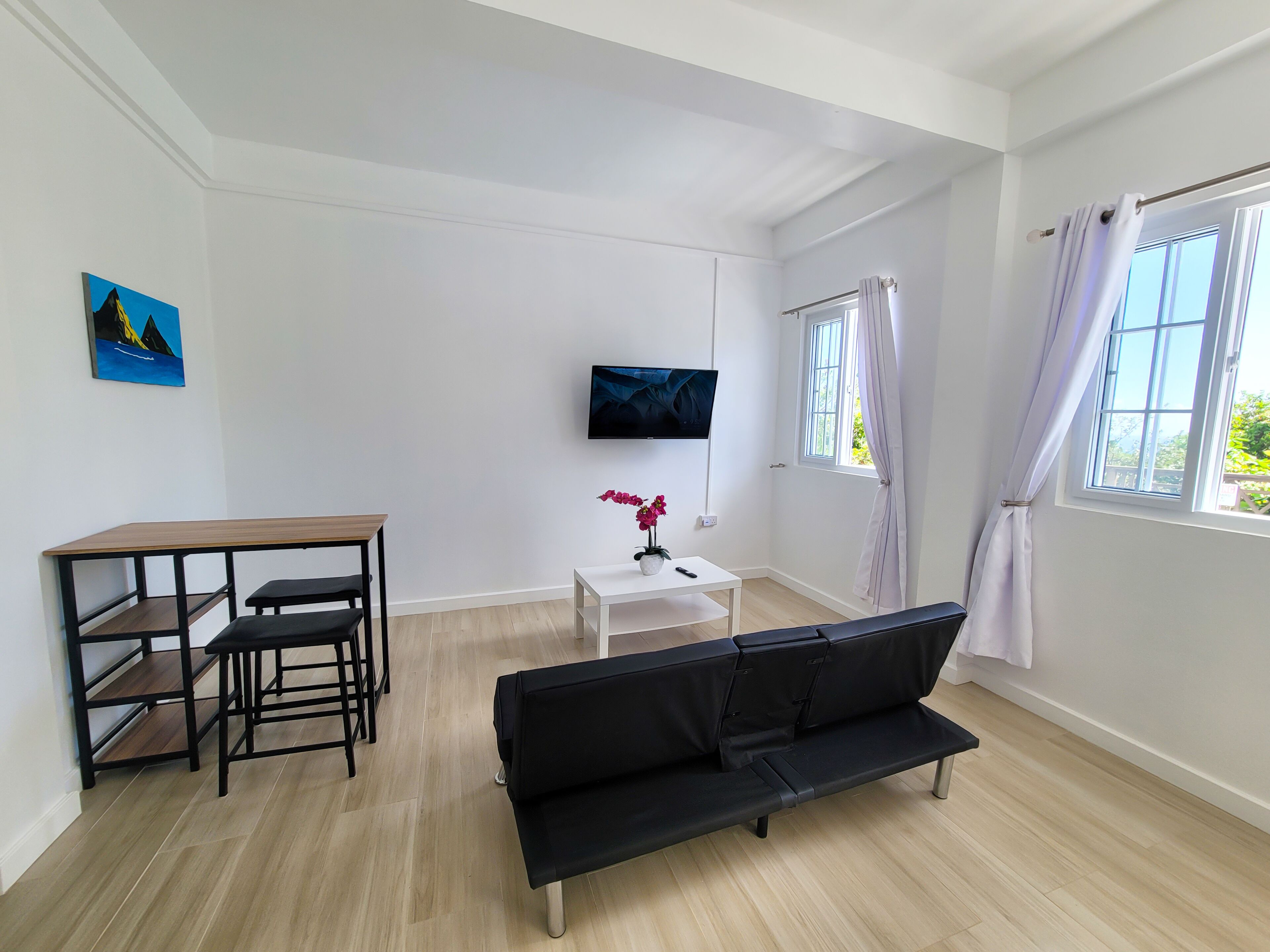 Un saló modern amb un sofà negre, una taula de dinar de fusta amb caires, un moble de televisió blanc amb un televisor de pantalla plana, i una finestra amb cortines.