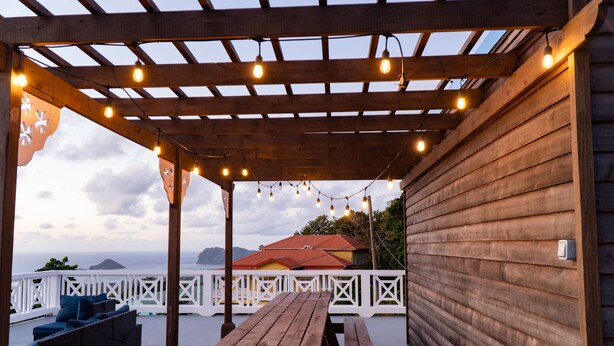 A wooden deck with a picnic table and wicker furniture, overlooking a scenic view with a wooden building and a red-roofed structure in the background. - Banbou Terrace (Vieux Fort)