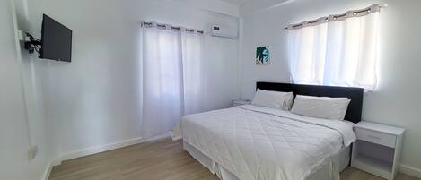 A bedroom with a bed, a nightstand, a wall-mounted TV, and a window with curtains.