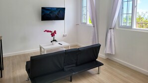 A modern room with a black sofa, a small white table with a vase of flowers, a mounted TV, and two windows with curtains. - Banbou Terrace (Vieux Fort)