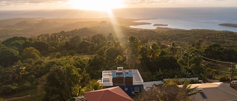 A house with a red roof, surrounded by lush greenery and palm trees, overlooking a coastal landscape with the ocean and hills in the distance.
