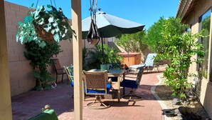 Un patio con una silla colgante, una mesa y sillas. Hay una planta colgante, una silla de madera y una pequeña mesa con un cojín azul.