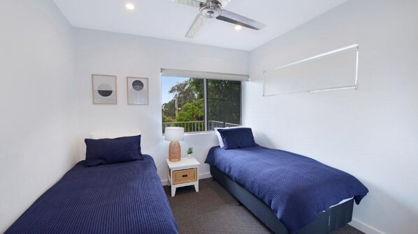 A bedroom with two beds, a window, a ceiling fan, and a small table with a lamp.
