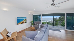 A spacious living room with a large gray sofa, a wooden chair, and a small table with a plant. A painting of a beach is on the wall, and there is a balcony with a view of the ocean.