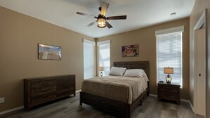 Ein Schlafzimmer mit Bett, Nachttisch, Kommode, Deckenventilator und zwei Fenstern mit Jalousien.
