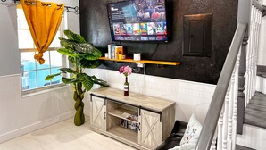 A living room with a TV mounted on a dark wall, a plant, a wooden cabinet, and a yellow curtain. - Amazing Ocean View Townhouse Condo Elevated Pool & Balcony (Nassau)