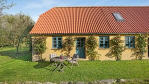 A yellow house with a red tiled roof, a front door, and two windows. There is a table and two chairs outside.