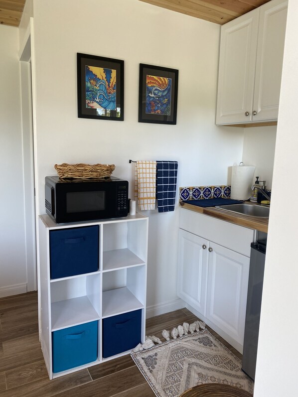 A compact kitchen with white cabinets, a microwave, and a sink. There are two framed artworks on the wall, a woven basket on the microwave, and a patterned rug on the floor.