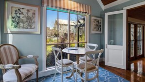 Un área de comedor con una mesa y sillas, una ventana que ofrece vistas a una casa y obras de arte enmarcadas en las paredes.