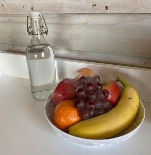 Un cuenco con fruta, incluyendo uvas, una manzana, una naranja y un plátano, sobre una superficie blanca junto a una botella de vidrio transparente.