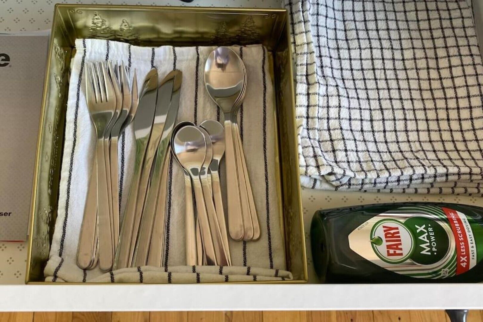 A drawer containing a set of silverware neatly arranged in a box, a green bottle of Fairmax powder, and a checkered cloth.