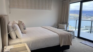 A modern hotel room with a large bed, a chair, a bedside table with a lamp, and a view of a lake through large windows. - Vues on Angelo | 3BR Home, Lake & Remarkables View (Queenstown)