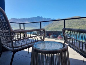 Un balcon agrémenté de fauteuils en osier et d’une table en verre, offrant une vue sur un paysage montagneux et une étendue d’eau.
