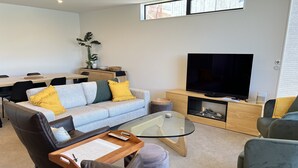 A modern living room with a sofa, coffee table, TV, and a dining area with chairs and a table. - Vues on Angelo | 3BR Home, Lake & Remarkables View (Queenstown)